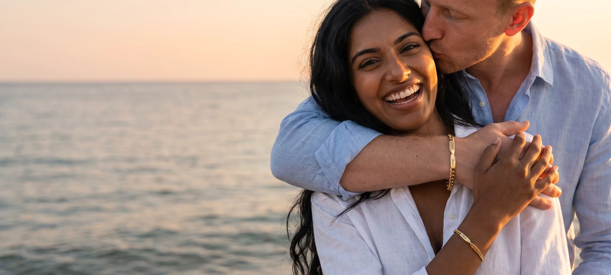 Un couple rit sur la plage au coucher du soleil, chacun porte son bracelet assorti en or